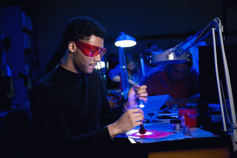 A Black male student wearing red glasses uses a flashlight to illuminate evidence in a crime lab lit by fluorescent black lights and a bright white lamp in the background. 